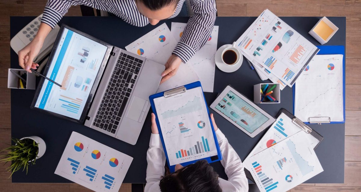 Equipe analisando gráficos de gestão em notebook, tablet e relatórios impressos